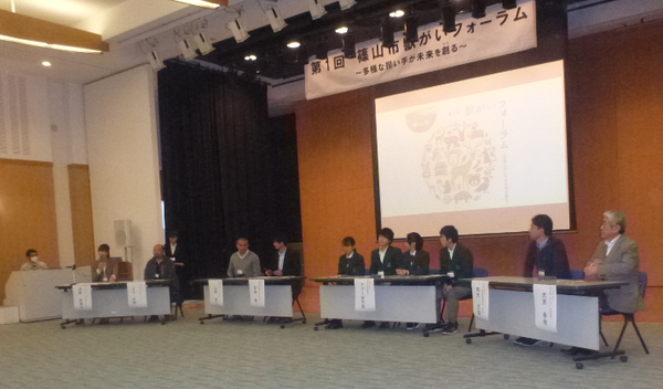 10名の大人や学生たちがモニターの前に座っていて、左はじの女性がマイクで話をしている写真