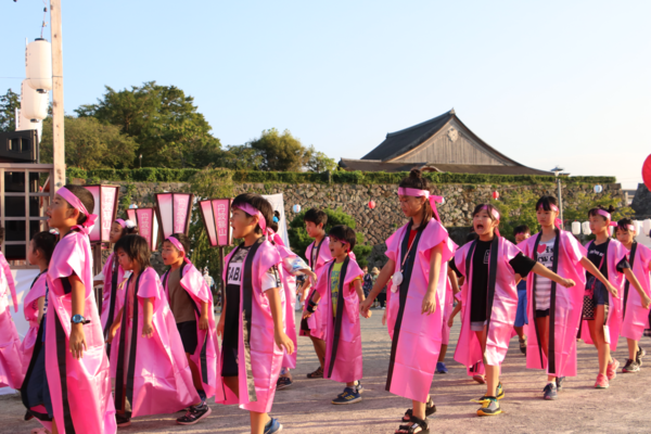 背丈の長いピンク色の法被を着た子供達が声を出してながら歩いている写真