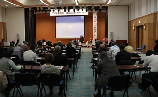 丹波篠山の景観ツーリズムと書かれた幕の横にモニターがあり、その前でマイクを持った男性が聴衆の方達に話をしている写真