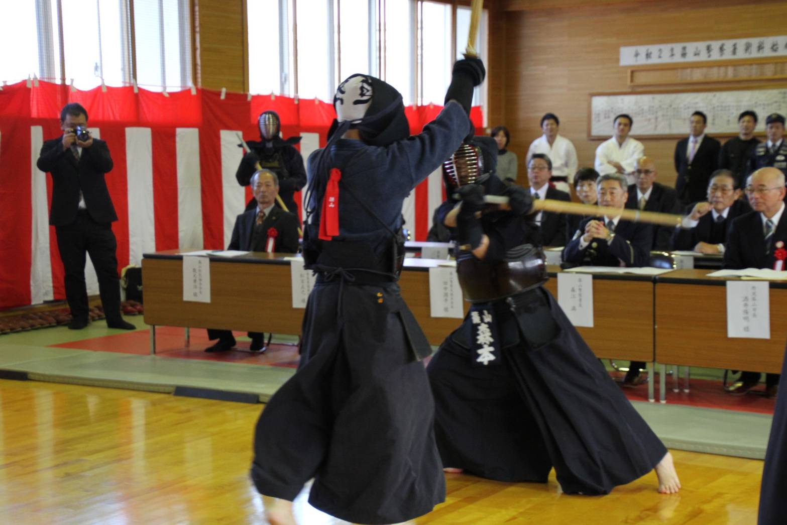 剣道の試合の様子と、それを見る術科始め式の出席者の人々の写真
