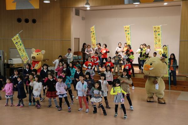 舞台では赤ちゃんを抱っこした女性たちが立ち、下では子供達がマスコット人形と踊っている写真