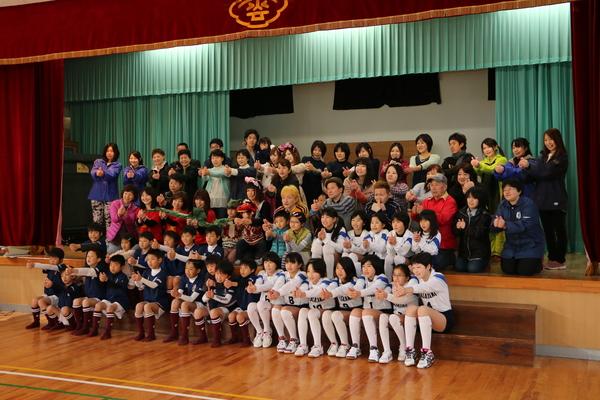 旧雲部小学校の体育館で、ユニフォームを着た生徒たちが両手でグーのポーズをして舞台の上にいる大人と一緒に記念写真