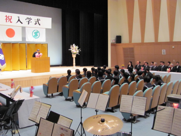 新入生、関係者の方々が、ステージ上で話をしている男性の方を向き、真剣に話を聞いている様子を左斜め後ろから写した写真