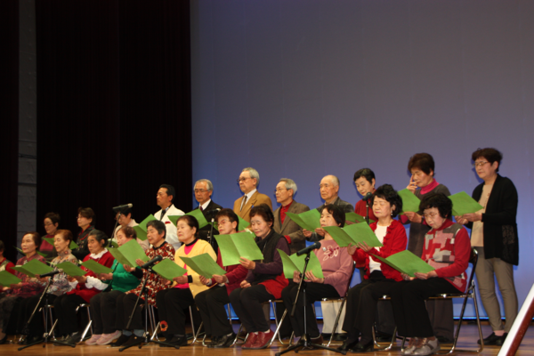 舞台の上で、緑の紙を見て2列に並ぶ年配の男女の写真