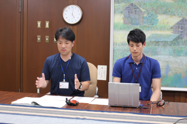 伊藤さんが机の上にパソコンを広げており、山本さんが活動報告の話をしている様子の写真