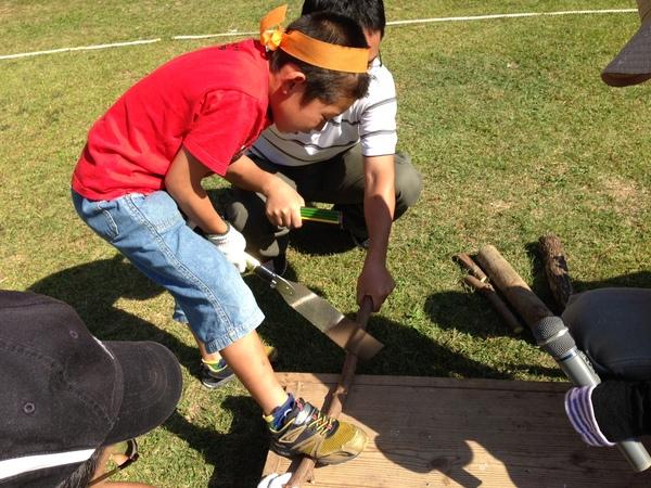 子供が一生懸命のこぎりで小さい丸太切って動かないように大人が支えている写真