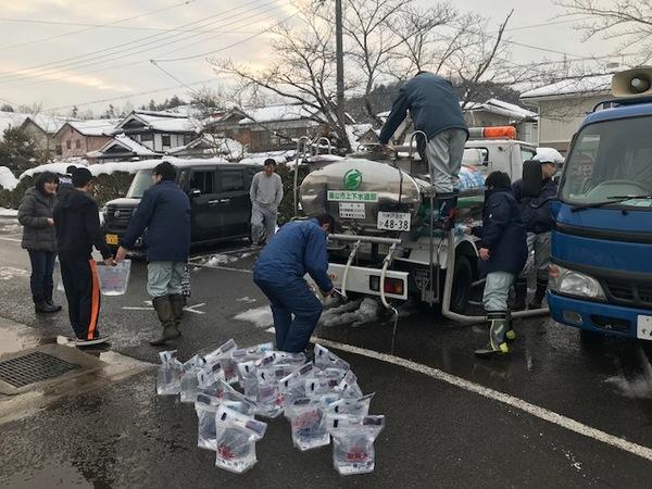 給水車から給水袋に水を入れて、それをもらいにきている人達と道路におかれた水がはいったたくさんの給水袋の写真