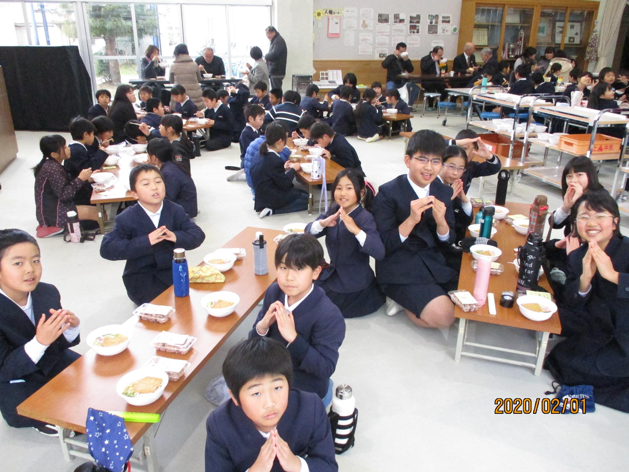 1つの長机の周りに5人ほどの生徒が座り、昼食を食べている写真。カメラを向けられている子どもたちは、両手の指の先を合わせて三角を作って写真に写っている。