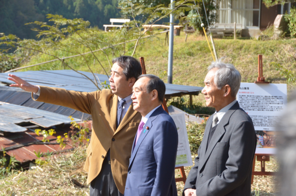 菅官房長官の両脇に職員が立っており右側の職員が手を伸ばして遠くを指さし、その先を菅官房長官と左側の職員が見ている写真