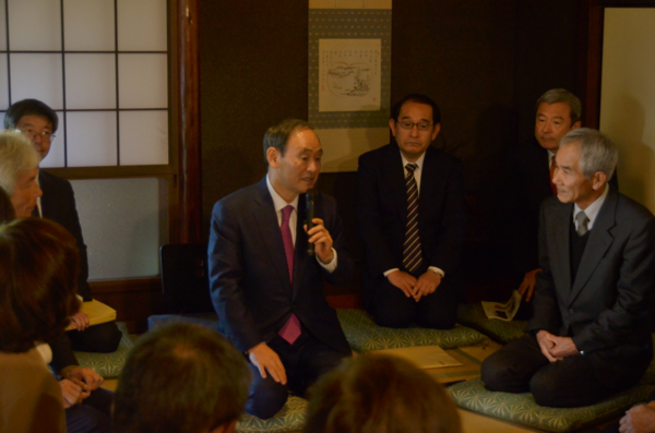 真ん中に菅官房長官が座ってマイクをもって話をしており、その周りを囲むようににスーツ姿の職員が座って話を聞いている写真