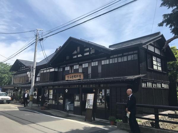 黒い大きなお店の前の隣に橋があり、橋の前で市長が立っている写真