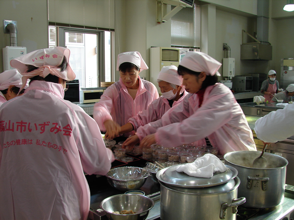 いずみ会と書かれたピンクのジャンパーを着て三角巾をつけた女性たちが調理をしている写真