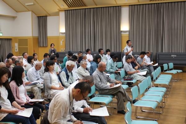 水色の椅子に参加者が腰かけ、資料を見ており、1人の男性がマイクを持ち、意見を述べている写真