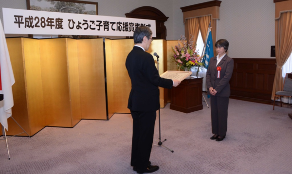 表彰式でスーツに赤い花を付けた女性が表彰をされている写真