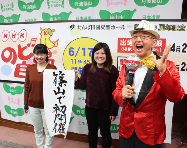 のど自慢の看板の前に、女性2人で篠山で初開催のチラシを持っていて、赤のジャケットに蝶ネクタイに手作りマイクを持った市長の写真