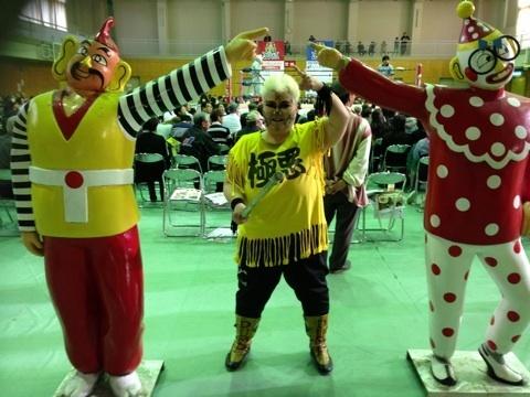 ピエロのような人形が右腕を上げて人差し指を突き出し、耳が大きく黄色い服にボーダー柄の袖の服をきた人形が左腕を上げ人差し指を突き出し、その間にダンプ松本さんが人形の人差し指に合わせるように左腕を上げて、人差し指を突き出し、ポーズをとって写っている写真