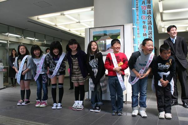 小学生の児童達が「すすんであいさつをしよう」のタスキを肩にかけて、元気よく挨拶をしている様子の写真