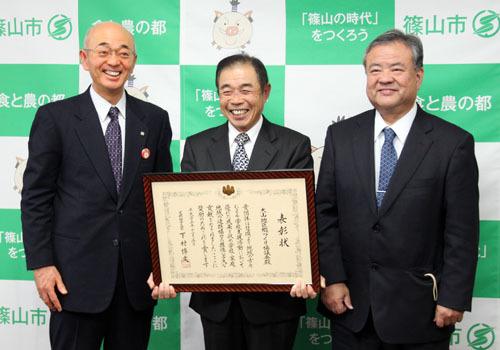 大山郷づくり協議会の斉藤会長が額に入った表彰状を手に持っており、市長と大山小学校の西山校長の3人が笑顔で写っている写真