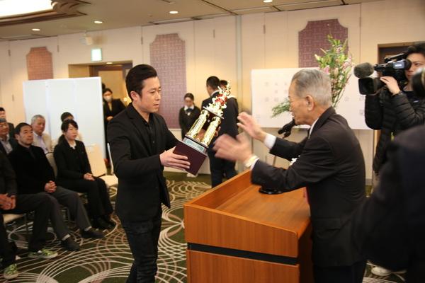 男性より大きなトロフィーを受け取る田中 久工さんの写真