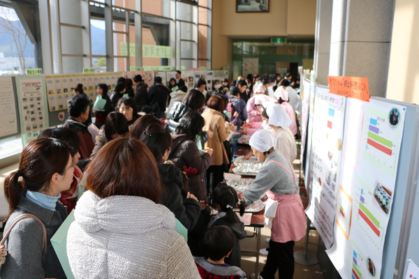 右側には三角巾をつけたエプロン姿の女性たちがテーブルに料理を置いて並んでいて、その前を大勢の人達が料理を見ながら歩いている写真