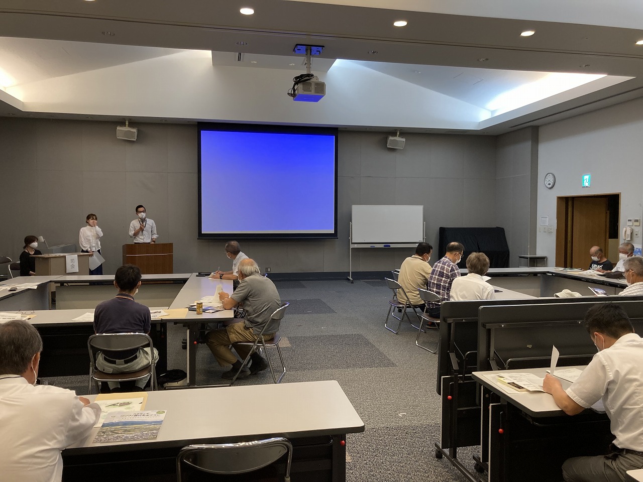 スクリーンの前で男性がマイクを持って話されていて、参加者が椅子に座って聞いている。