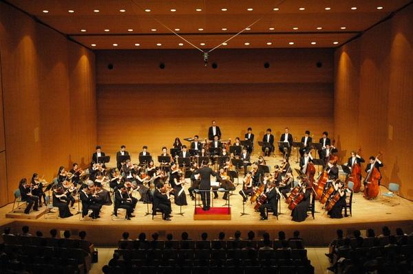 舞台で黒い服装をした管弦楽団が指揮者に合わせて演奏しているのを観客席で鑑賞している人々の写真