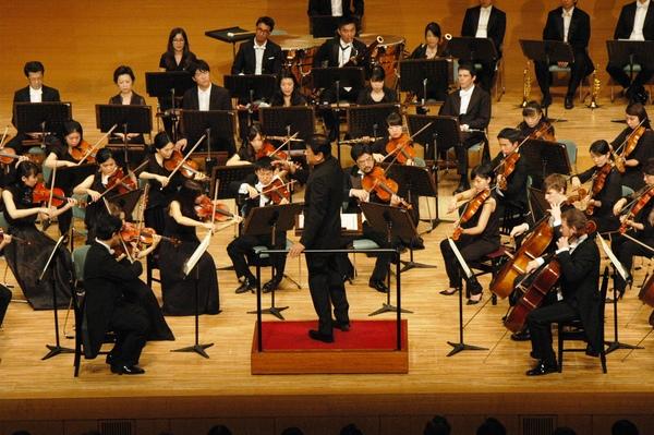 舞台で黒い服装をした管弦楽団が指揮者に合わせて演奏している写真