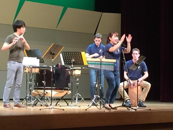 打楽器とフルートの演奏をバックにスタンドマイクで女性が歌っている写真