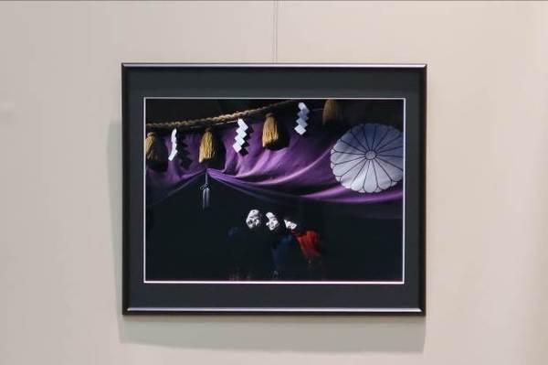 紫色の幕に菊花紋章が入っており、その前にしめ縄があり、紫の幕の下から、白いおかめのお面を被った3人の人が顔のぞかせている写真で「節分の日」という題名の作品の写真
