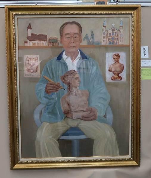 メガネを掛け、青藍色のジャンパーを着た古希を過ぎた男性が、椅子に座っており、右手に彫刻刀を持ち、左手で女性の上半身の木彫りの置物を持っている様子が描かれた絵画の作品の写真