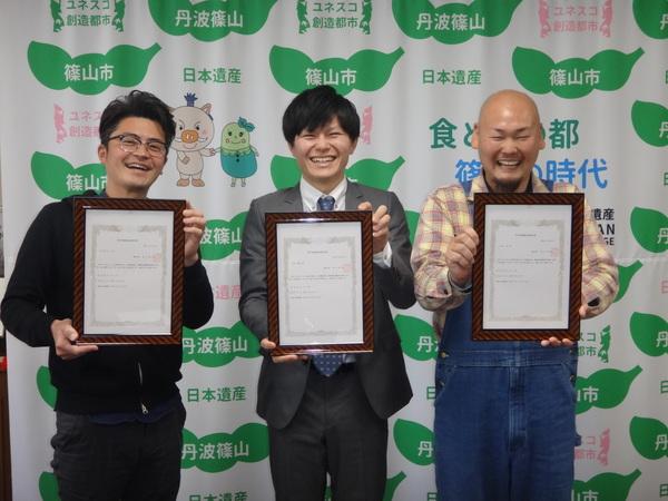 市から交付された「青年等就農計画認定書」を手に持った笑顔の男性3名が写っている記念写真
