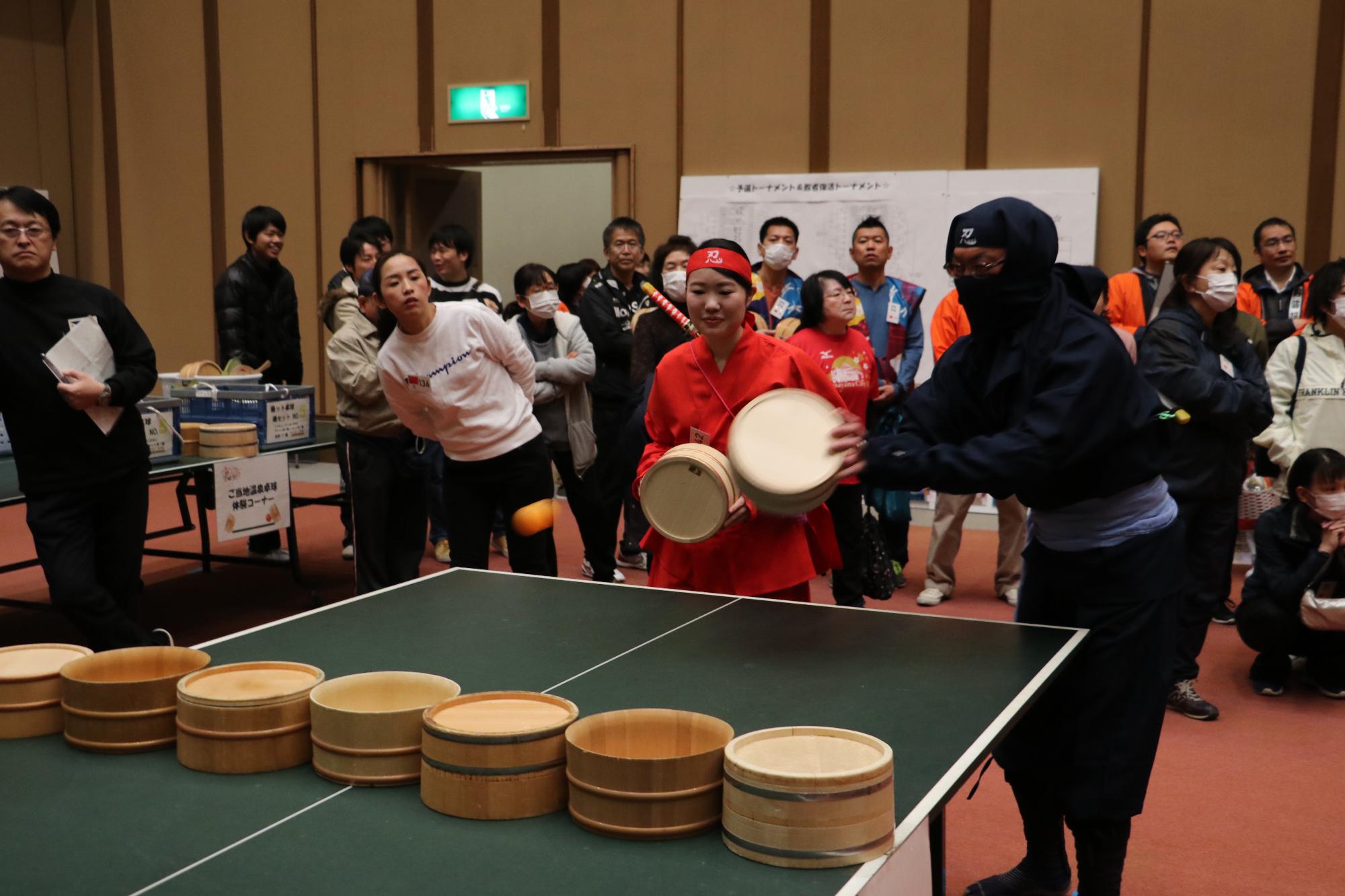 一人は赤色、もう一人は黒色の忍者の格好をしたペアがピンポン玉を打ち返している写真