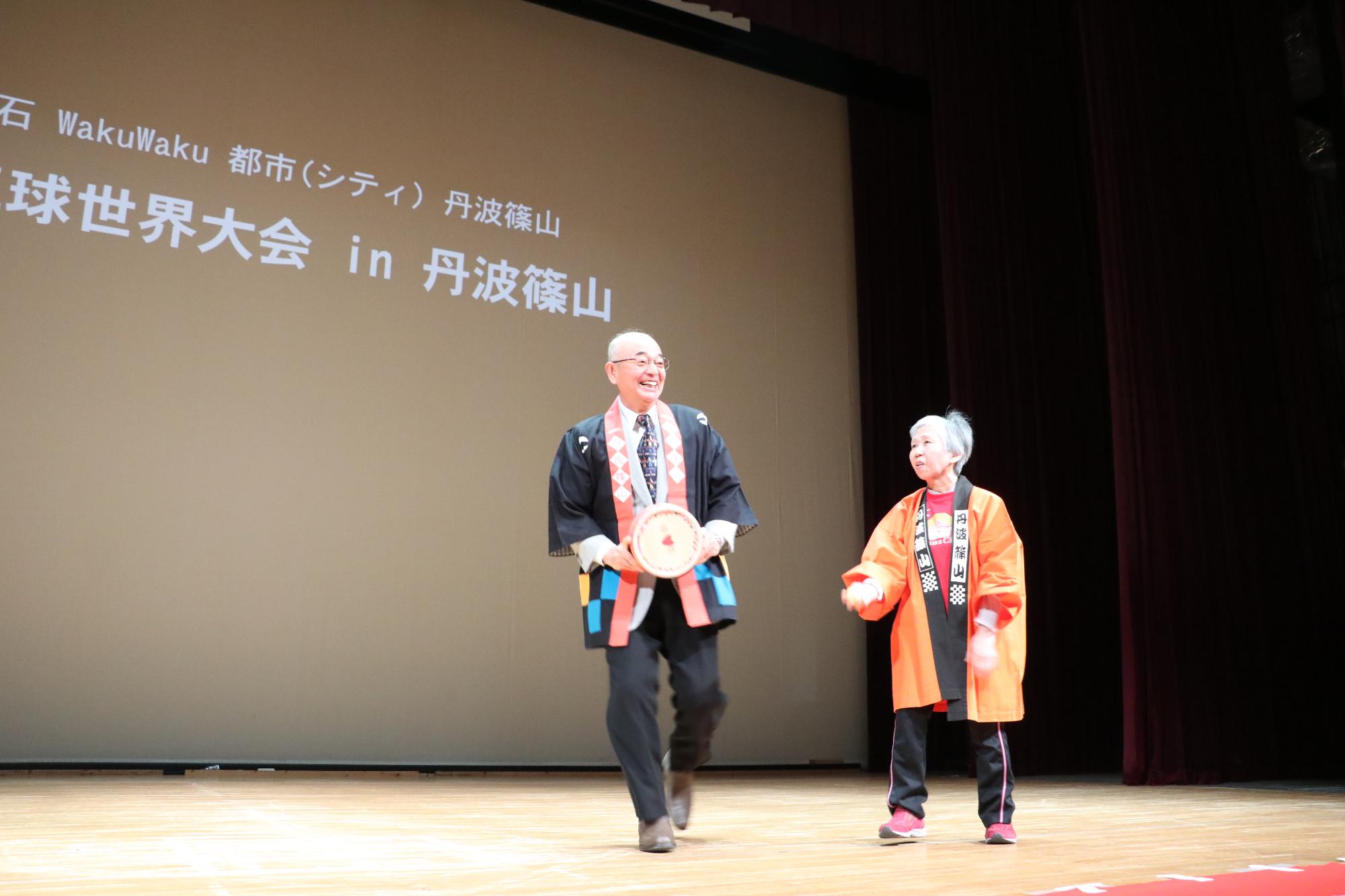 黒いハッピを着た笑顔の酒井市長が桶を両手で持って舞台上にいる横でオレンジ色の丹波篠山と書かれたハッピを着ている女性が立っている写真