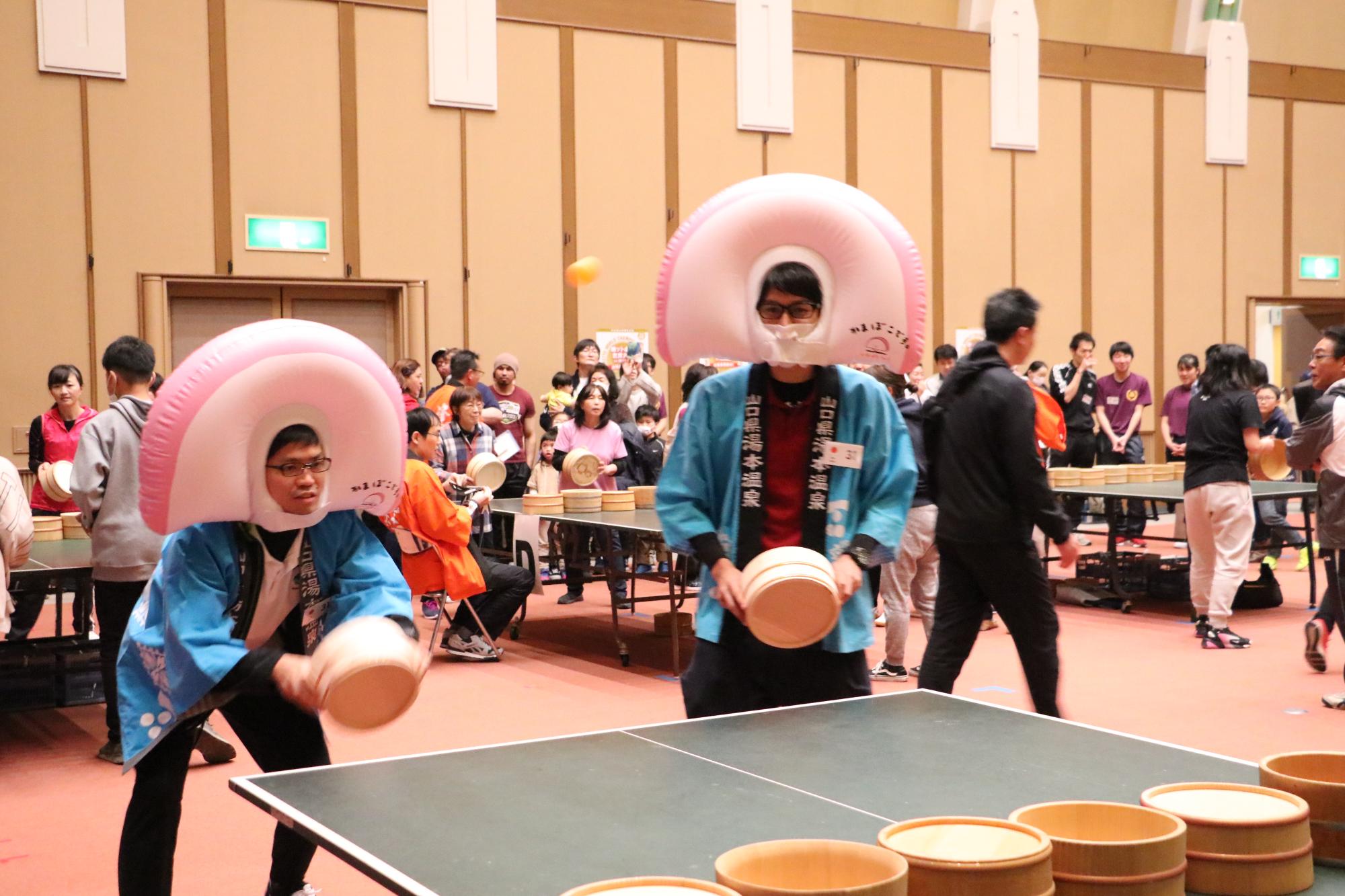 山口県湯本温泉と書かれた青いハッピと、半円の形をしたかまぼこの被り物をかぶった男性2人がペアになって試合をしている写真