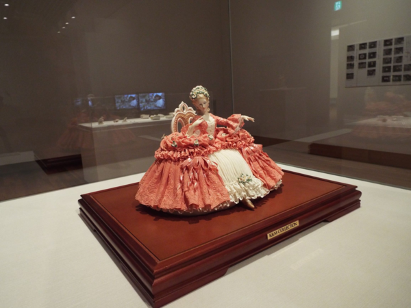ショーガラスの中の、椅子に座るオレンジ色のドレスを着た人形の置物の写真
