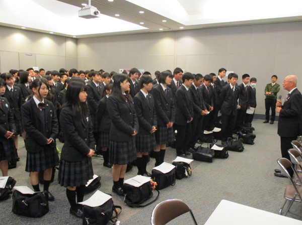 高校生が足元にカバンを置き起立して市長の話を聞いている写真