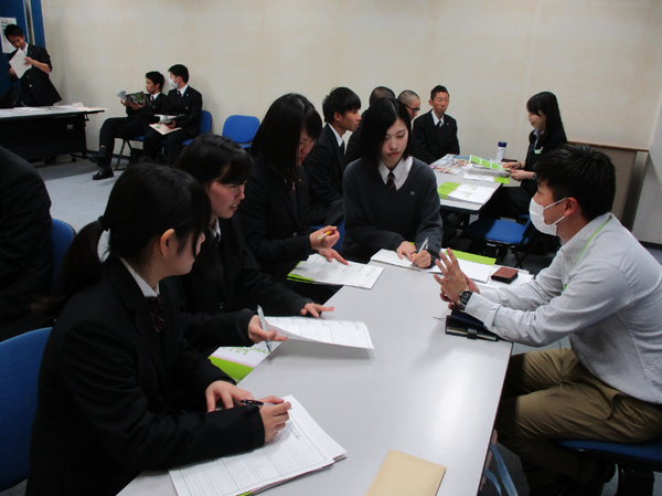 マスクをした企業の男性が女子高生4人に手を使い説明している写真