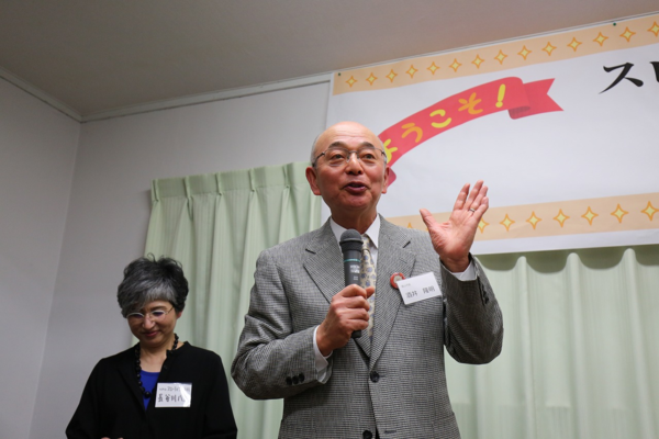 市長がマイクを持ってお話ししている後ろで眼鏡をかけられた女性が静かに聞いている写真