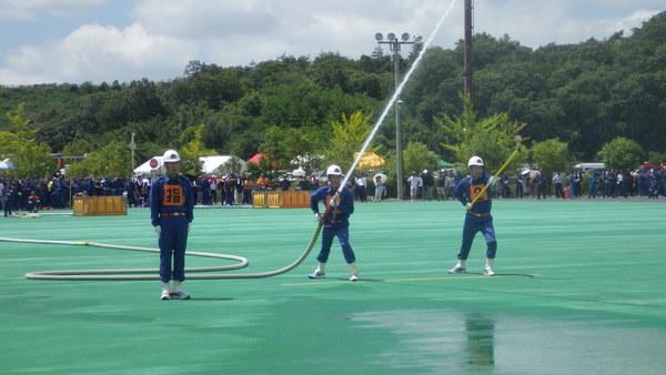 胸に、番号と指と書かれたユニフォームを着た男性が、ホースを持ち放水している写真