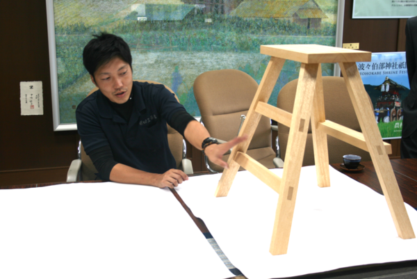 机の上に置かれた、四方（しほう）転び踏み台の脚の部分に手を添えて園田さんが説明している様子の写真