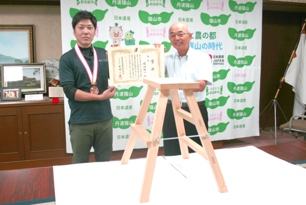 四方（しほう）転び踏み台を机の上に置き、その後ろに園田 雄一さんが首からメダルを下げて賞状を市長と一緒にもって写っている写真