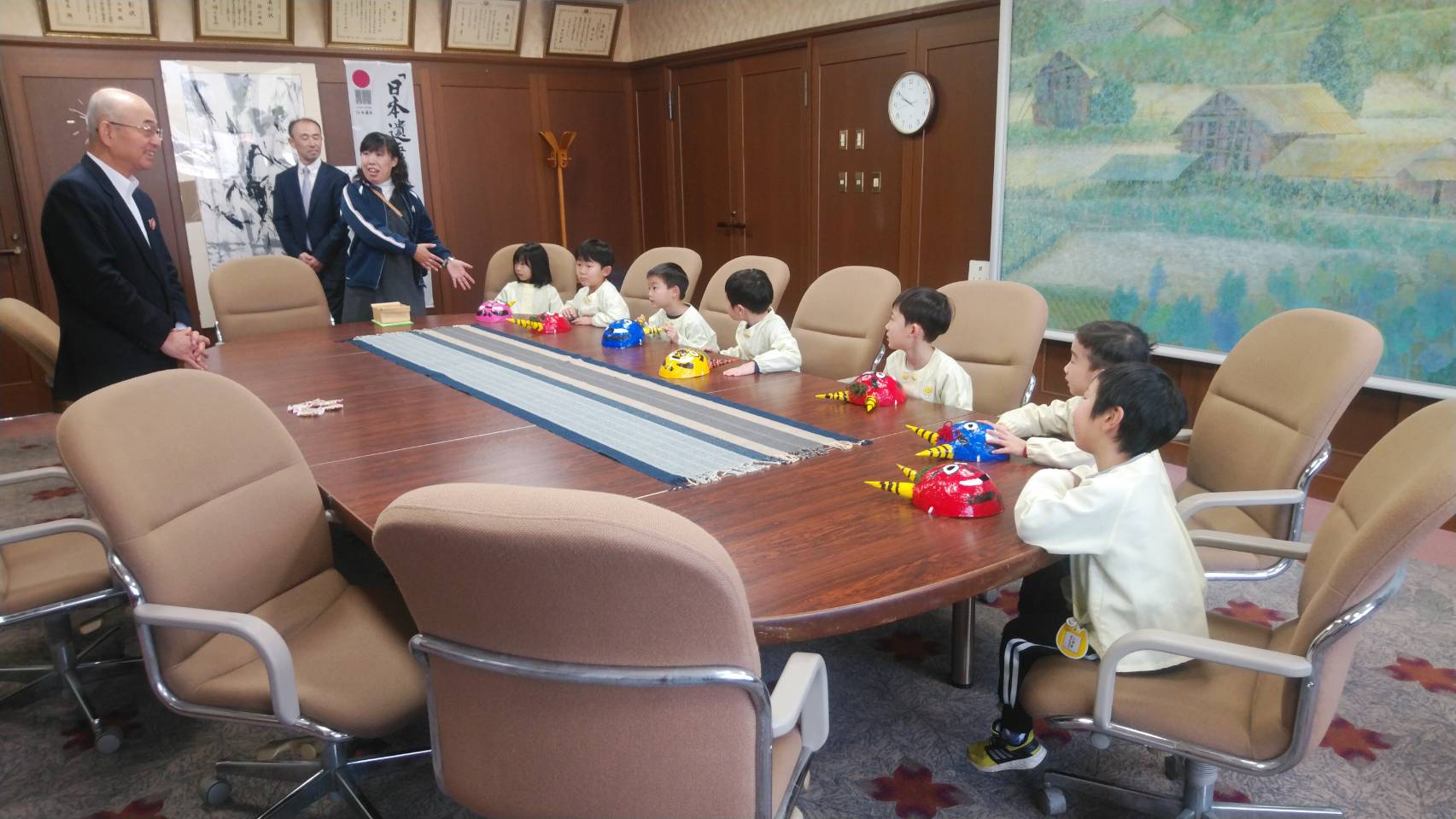 色とりどりの鬼のマスクを机の上において応接室のイスに座る幼稚園児のみなさんと、幼稚園の先生と、市長の写真