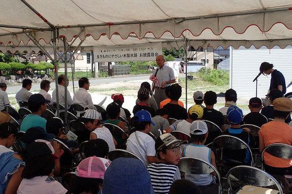 木製水路のお披露目会で、テントの中に座っている沢山の子供たちと、関係者、その前で話す市長の写真