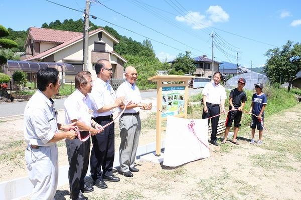 木製水路の横に作られた、看板の前で、関係者と市長、男の子2人が一緒にリボンを持っている写真