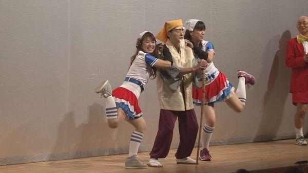 舞台に杖を突いて立っている黄門さまを真ん中にチアガール風の衣装を着て日本タオルを頭にのせた女性が黄門さまに寄り添って片足を上げて立っているのを市長が見ている写真