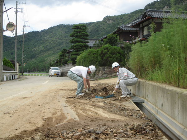 道路沿いの側溝の泥やがれきが道路に置かれており、職員の男性2名が片付けをしている様子の写真