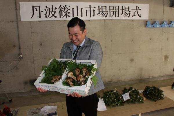 丹波篠山市場開場式と書かれた紙の貼られた壁の前で男性が松茸の箱を2箱持って笑っている写真