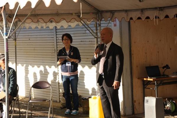 テントの下でスーツを着た男性がマイクを持って話をしている写真