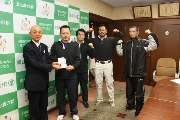 代表の西野 裕彦さんと市長が奨励金を持ち、後ろの3名の選手がガッツポーズをしている写真