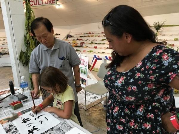 市野先生が外国人の女の子に習字を教えている様子を隣で女性がみている写真